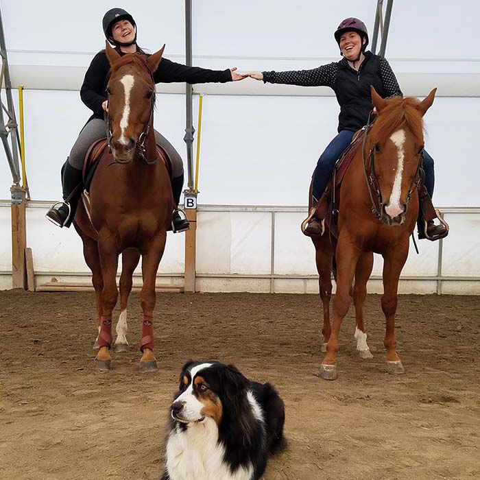 Two women on two separate horses holding hands, with a dog in the middle Two women on two separate horses holding hands, with a dog in the middle
